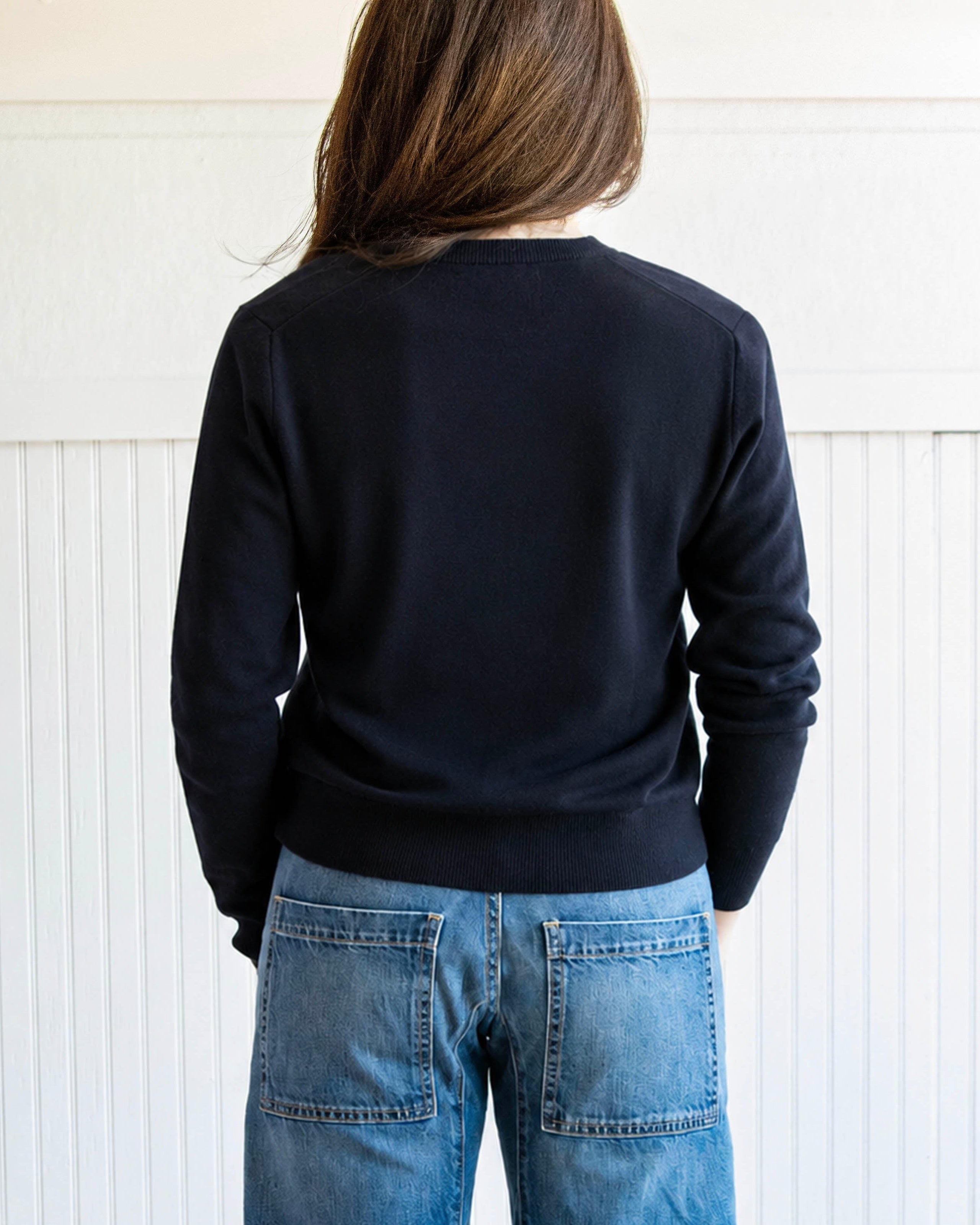 Person wearing a black sweater and blue jeans standing against a white paneled wall.